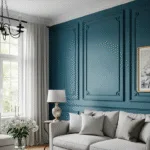 Elegant living room with a blue paneled accent wall, light gray sofa with matching pillows, round side table with a lamp, white curtains, window, framed artwork, and a vase of white flowers on a small table.