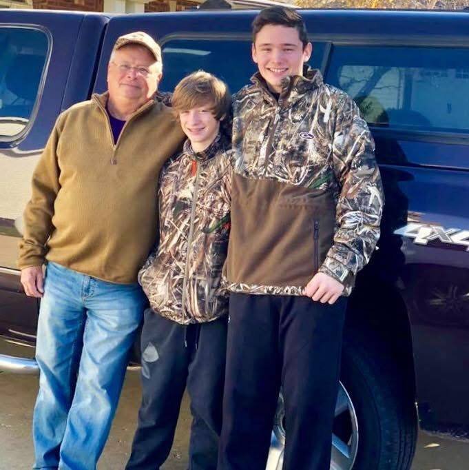Three generations of men stand in front of a truck, wearing casual and hunting attire.