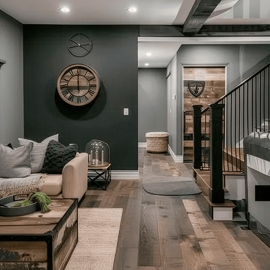 A modern living room with dark walls finished by residential painting Orlando, a beige sofa, a large rustic clock, decorative pillows, a wooden coffee table, and a staircase with black railings. The space features wood flooring and cozy, neutral decor.