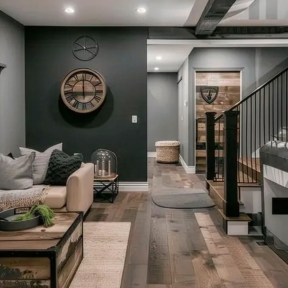 A modern living room with dark walls finished by residential painting Orlando, a beige sofa, a large rustic clock, decorative pillows, a wooden coffee table, and a staircase with black railings. The space features wood flooring and cozy, neutral decor.