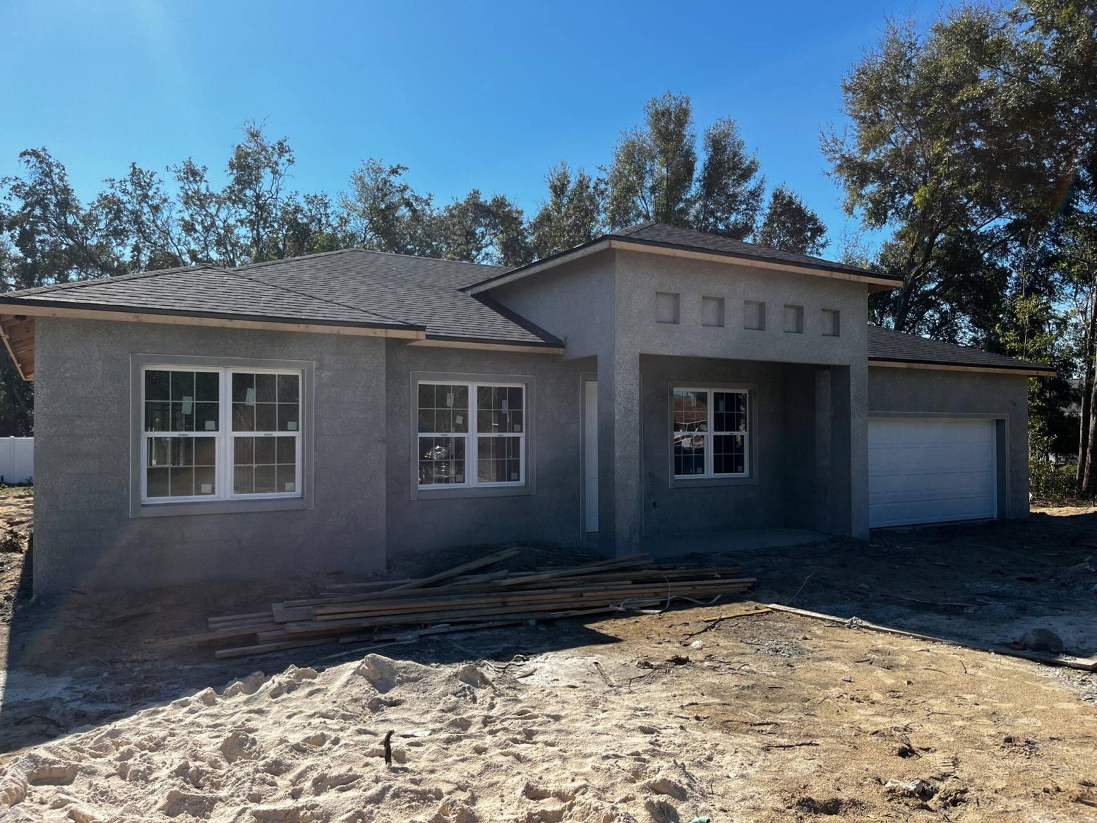 New construction home with gray stucco exterior, dark roof, and white windows under a blue sky.